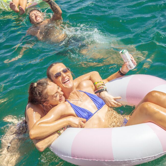 Two women in swimsuits laugh and embrace on a pink-and-white pool float in bright blue water, while a person swims and smiles in the background. One woman holds a beverage can. Sunlight reflects off the water.