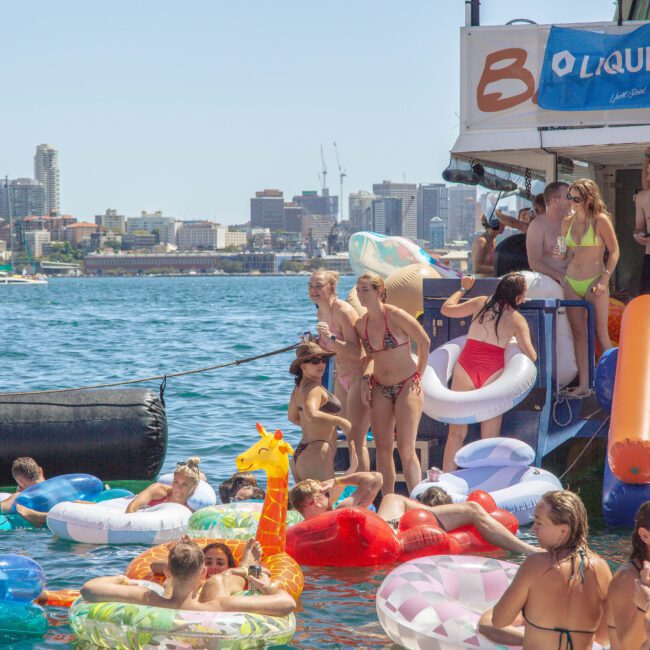 A lively group of people in swimsuits enjoy a floating party with colorful inflatable tubes and animal floats on the water, near a docked boat, with a city skyline in the background.