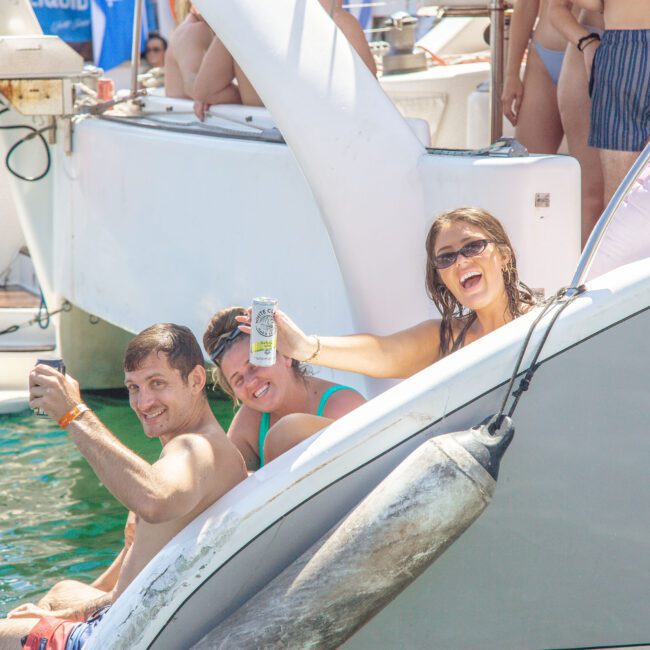 Three smiling people in swimsuits sit on the edge of a yacht, holding drinks and posing for a photo. Other people and boats are visible in the background, suggesting a lively summer gathering on the water.