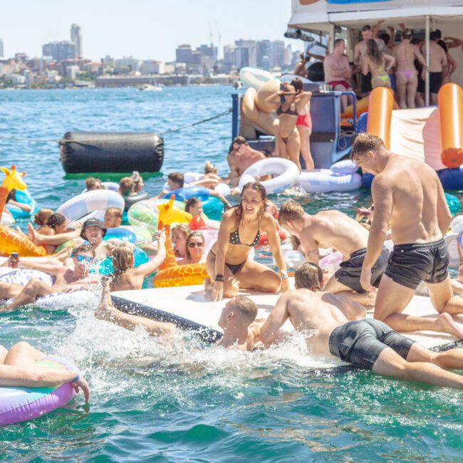 A lively scene of people enjoying a sunny day on the water, floating on inflatable rafts and pool toys near a crowded boat, with a city skyline visible in the background.