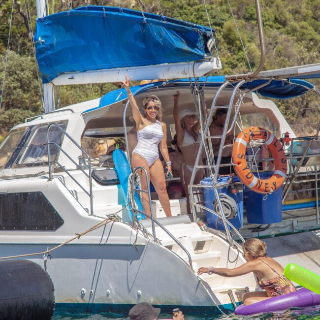 A woman in a white swimsuit waves from a yacht deck, while others relax nearby in swimsuits. The yacht is anchored by a lush, green shoreline, with inflatables floating in the water.