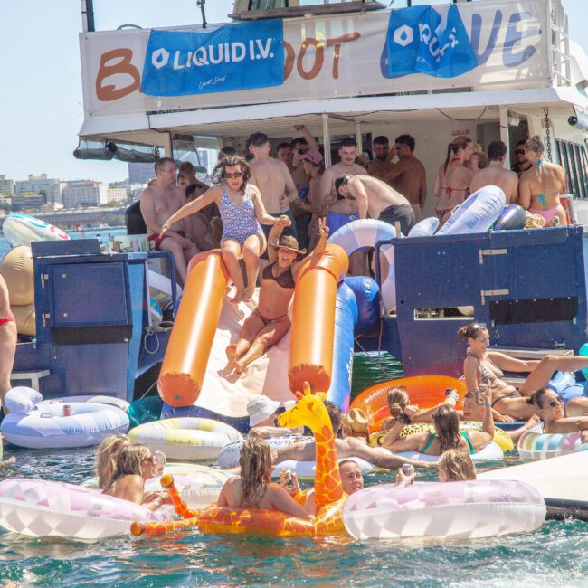 A group of people enjoy a lively party on a boat and in the water, surrounded by colorful pool floats, including a giraffe float. Some people use orange slides to enter the water. The city skyline is visible in the background.