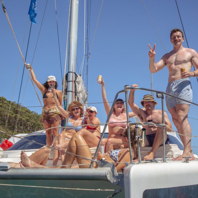 A group of people in swimwear enjoy drinks and pose cheerfully on the deck of a sailboat under sunny skies, with trees visible in the background. One man makes a peace sign and others smile at the camera.