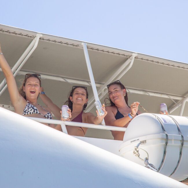 Three women in swimsuits smile and wave from the deck of a boat, each holding a drink. A white life raft is mounted on the railing nearby, and the sky is clear and blue.