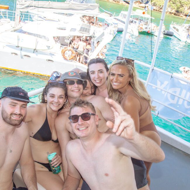 A group of six smiling young adults in swimsuits pose together on a boat, with turquoise water and other party boats crowded with people visible in the background on a sunny day.