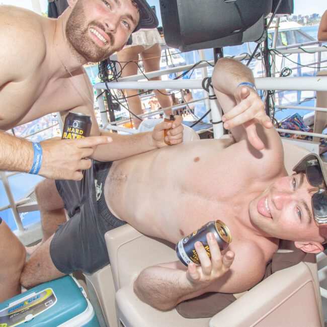 Two young men in swimwear pose playfully with canned drinks at a boat party. One is sitting and sticking out his tongue while pointing; the other stands next to him, smiling. People and boats are visible in the background.