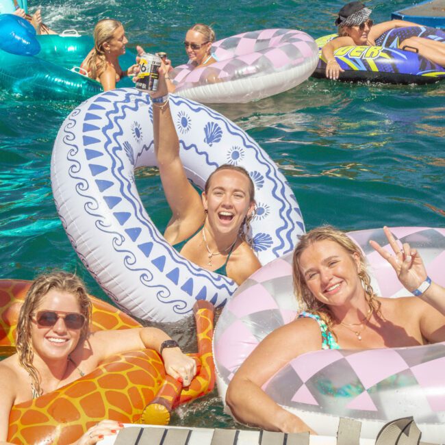 A group of smiling people float on colorful inflatable tubes in a pool, with two women in the front making peace signs and another raising her arm in excitement. The scene is bright and lively.