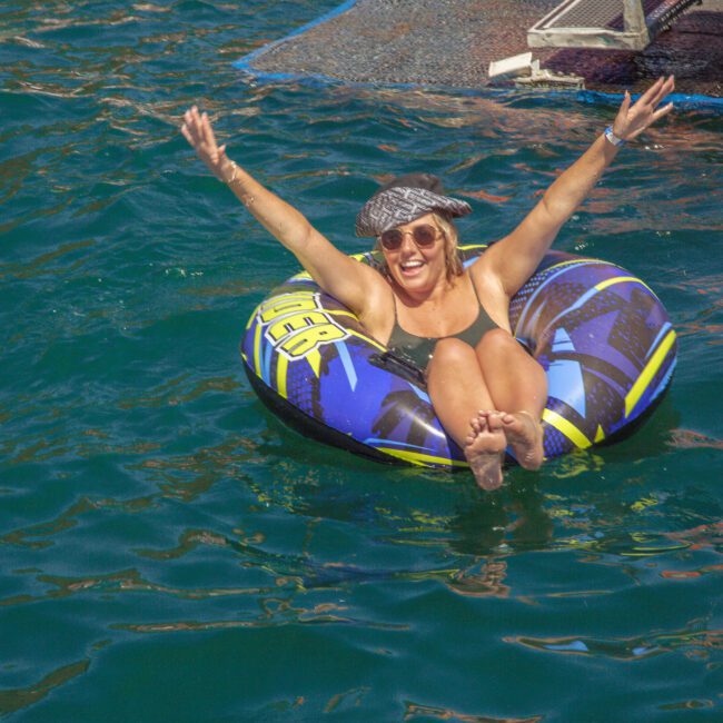 A woman in sunglasses and a hat smiles with her arms raised while floating on an inflatable tube in the water near a dock. The sun is shining and the water is a vibrant green-blue.