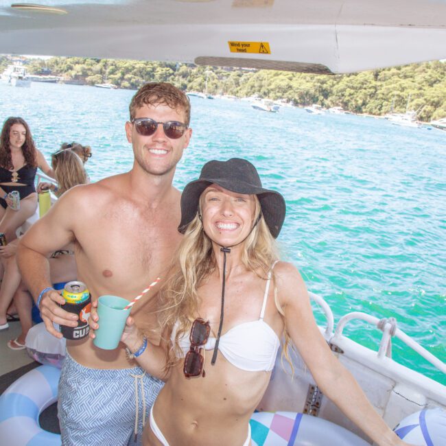 A smiling man and woman in swimwear pose together on a boat. The woman wears a sunhat and white bikini, while the man wears sunglasses and blue swim trunks. Blue water and trees are visible in the background. Other people are on the boat.