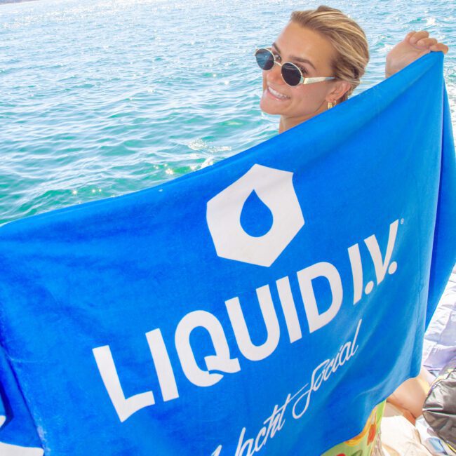 A person wearing sunglasses smiles and holds a bright blue towel that says "LIQUID I.V. Yacht Social" by the water, with the Sydney Harbour Bridge visible in the background.