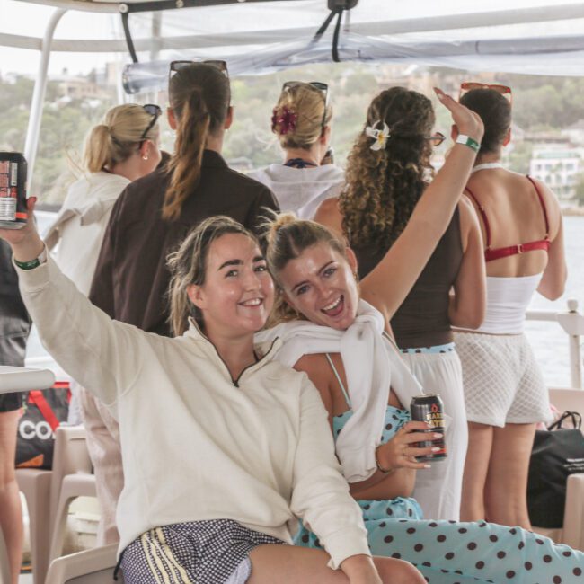 Two women smiling and raising drinks while sitting on a boat; people in summer outfits stand in the background, facing the water. The scene looks lively and relaxed, with a coastal view visible outside.