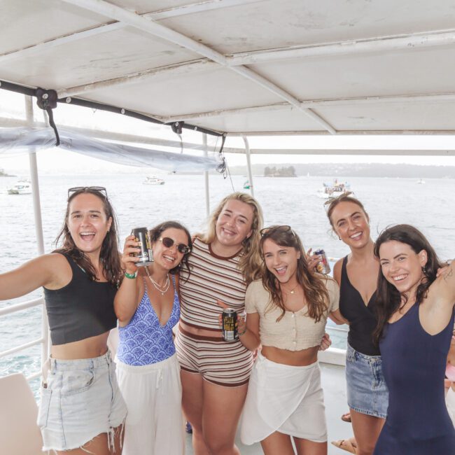 Six smiling women stand on a boat holding drinks, posing for a cheerful group photo with the ocean and other boats visible in the background. Some are wearing swimsuits and summer clothes.