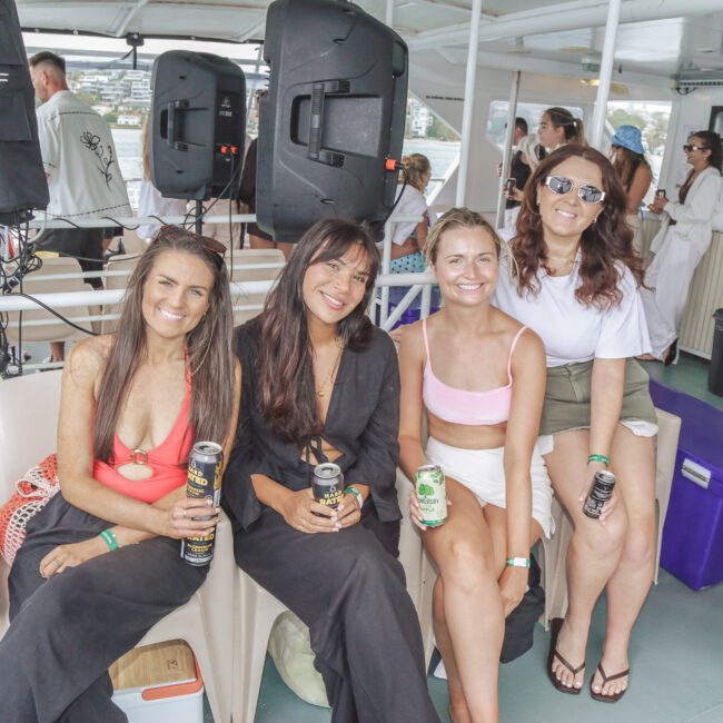 Four women sit smiling on a boat, holding drinks and wearing summer outfits. Two are in black dresses, one in a coral bikini top, and one in a white crop top. People and large speakers are in the background.