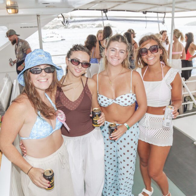 Four smiling women in swimwear and sunglasses pose together on a boat, each holding a drink. The background shows other people, the boat’s interior, and water outside. The mood is relaxed and festive.