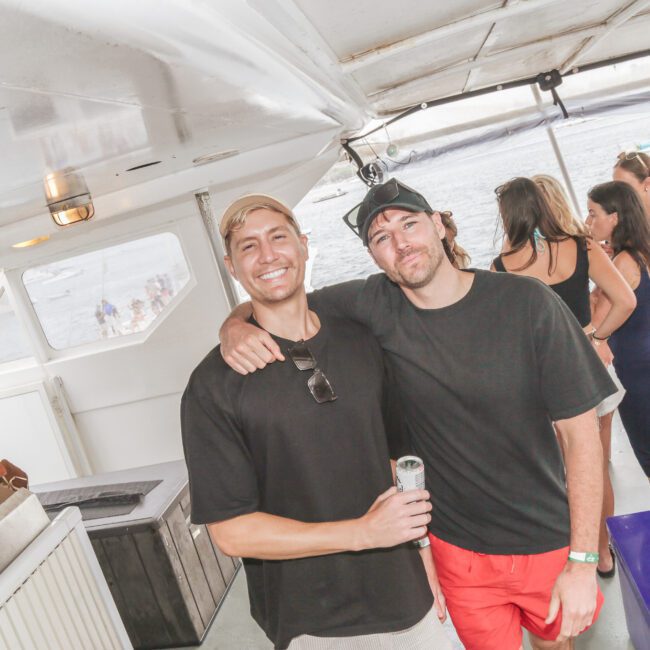 Two men pose and smile on a boat; one has his arm around the other. Other people are in the background, and boats are visible on the water outside. The scene appears casual and relaxed.
