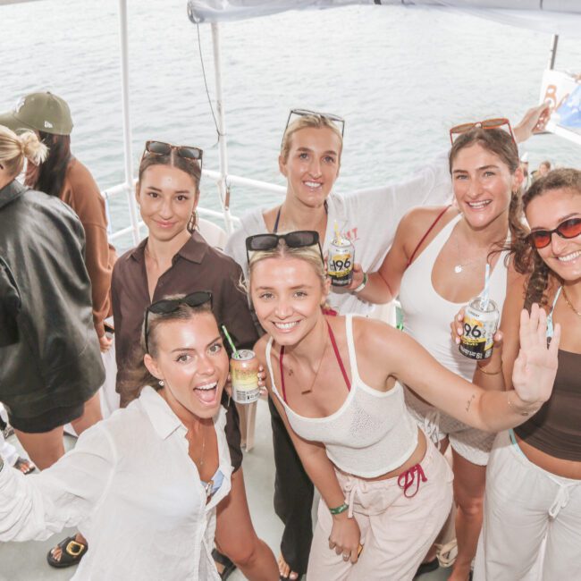 A group of six smiling women pose together on a boat, holding drinks and enjoying a sunny day. Other people are visible in the background, and the sea can be seen beyond the deck.