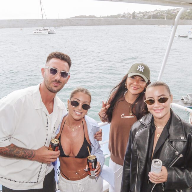 Four people posing and smiling on a boat, holding drinks. The group includes two women in sunglasses, one in a black jacket and one in a bikini top, plus a man in a white shirt and a woman in a brown sweatshirt and green cap. Boats and water are in the background.