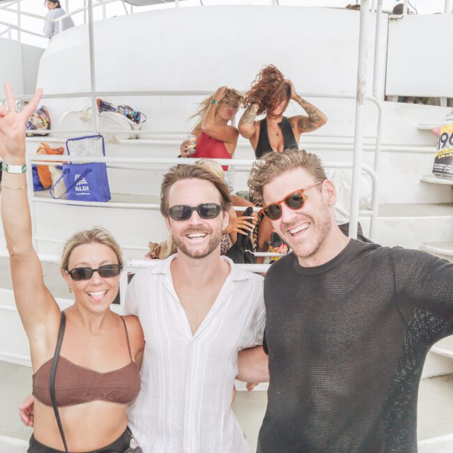 Three people pose and smile on a boat, with one woman holding up a peace sign and sticking out her tongue. Two men stand beside her, one holding a can. Several others sit and relax in the background. The mood is joyful and casual.