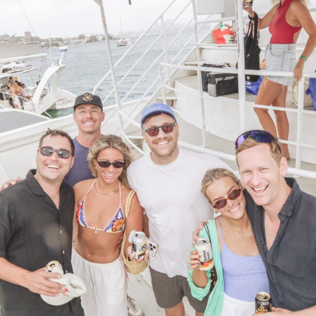 A group of six smiling people stand together on a boat, holding drinks and wearing sunglasses and summer clothes, with water and another boat visible in the background on a cloudy day.