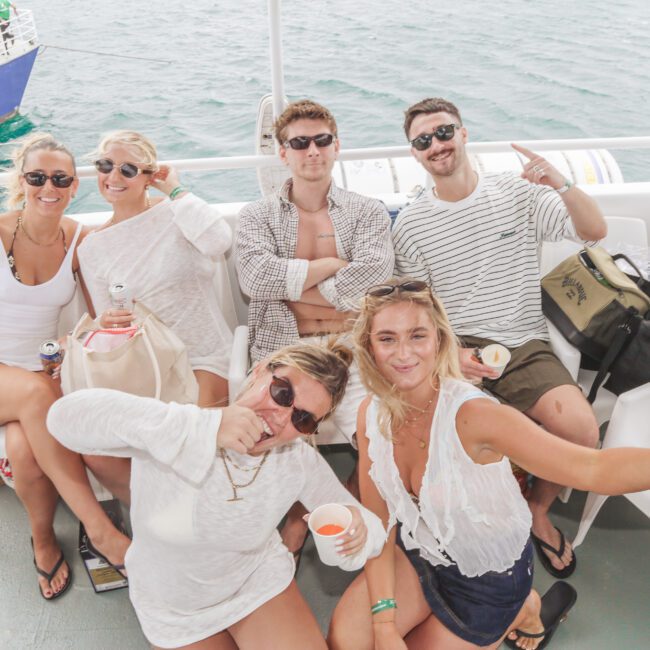 Six young adults in casual summer clothes sit and smile on a boat, posing for the camera. The ocean and another boat are visible in the background. Some hold drinks and snacks, enjoying a relaxed, sunny outing.