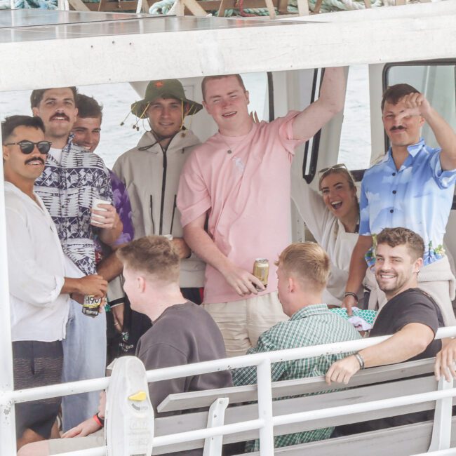 A group of people, mostly men, sit and stand on a boat, smiling and waving at the camera. Some are holding drinks and wearing casual summer clothes. The boat is on the water, and it appears to be a social gathering.