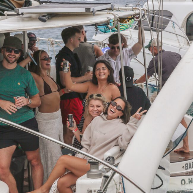 A group of smiling people relax, laugh, and pose for a photo on a boat, holding drinks. The boat is docked near other boats on the water and everyone appears to be enjoying a fun, casual outing.