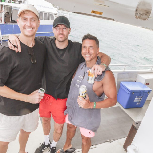 Three men stand closely together smiling on a boat, each holding a drink. The ocean and another boat are visible in the background. One man wears black shorts, another wears red shorts, and the third wears pink shorts.