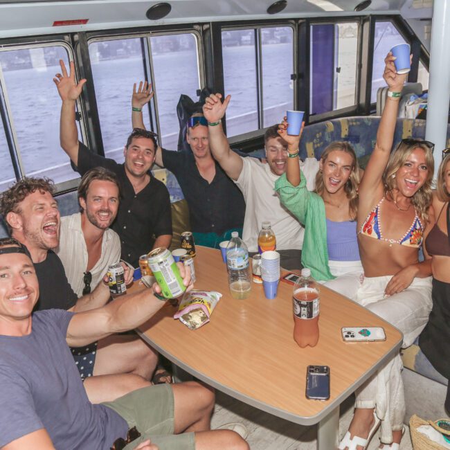 A group of nine smiling people sit around a table on a boat, raising drinks and snacks in celebration. The ocean is visible through the windows behind them. The atmosphere is festive and relaxed.
