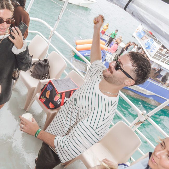 A group of people are enjoying a party on a boat. One man in sunglasses and a striped shirt raises his arm while holding a drink, with others around him smiling and talking. The ocean and other boats are visible in the background.
