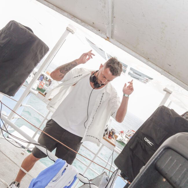 A DJ wearing headphones and a white shirt is performing on a boat, surrounded by speakers and equipment, with water and other boats visible in the background. The scene appears lively and energetic.