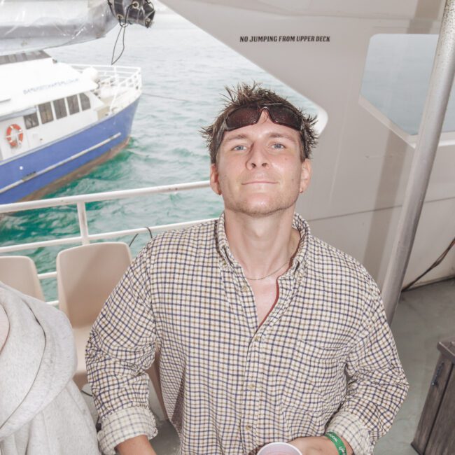 A man in a plaid shirt stands on a boat holding a red cup, looking up at the camera. Another person is partially visible to his left. A blue and white boat is seen in the water outside. A sign reads "NO JUMPING FROM UPPER DECK.