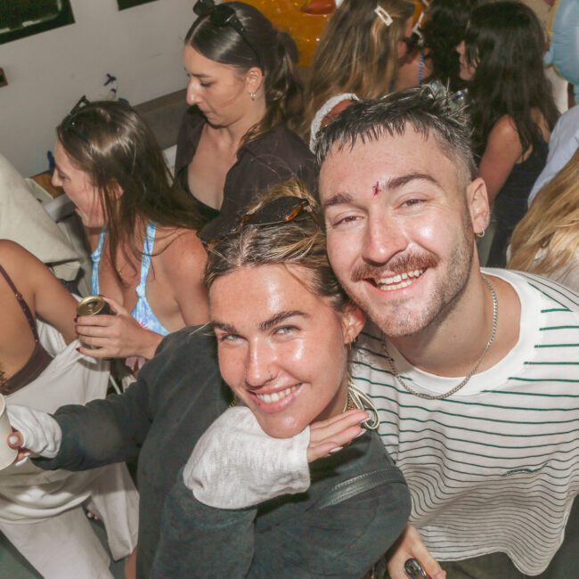 A group of young adults at a lively indoor party. Two people in the foreground smile at the camera, with others chatting and mingling behind them. The atmosphere is energetic and celebratory.