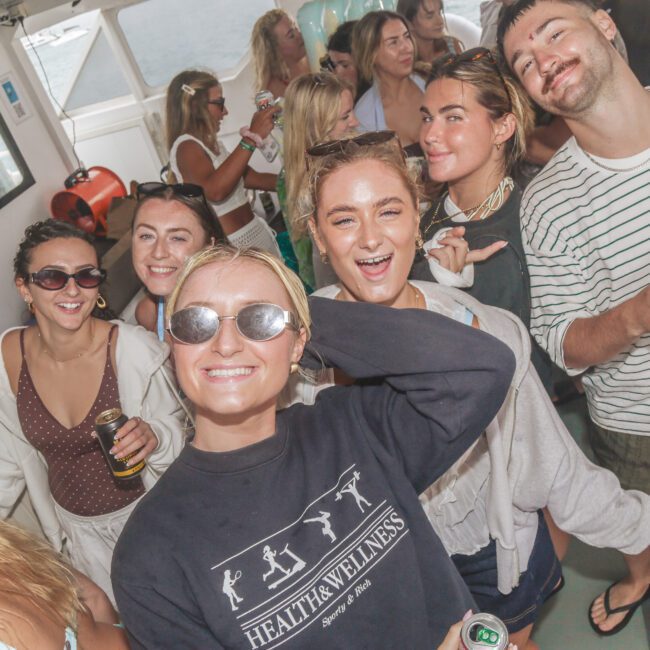 A group of young adults smile and pose for a photo at a lively party on a boat. Some people wear sunglasses and hold drinks, while others laugh and enjoy themselves in the sunny, casual atmosphere.