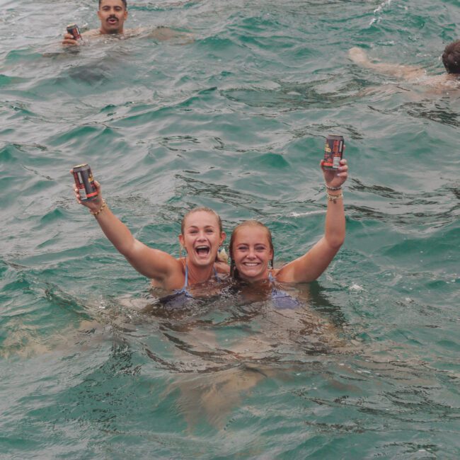 Two women smiling and holding up canned drinks while swimming in the ocean, with other people swimming in the background. The water is a vibrant blue-green, and the mood is joyful and celebratory.