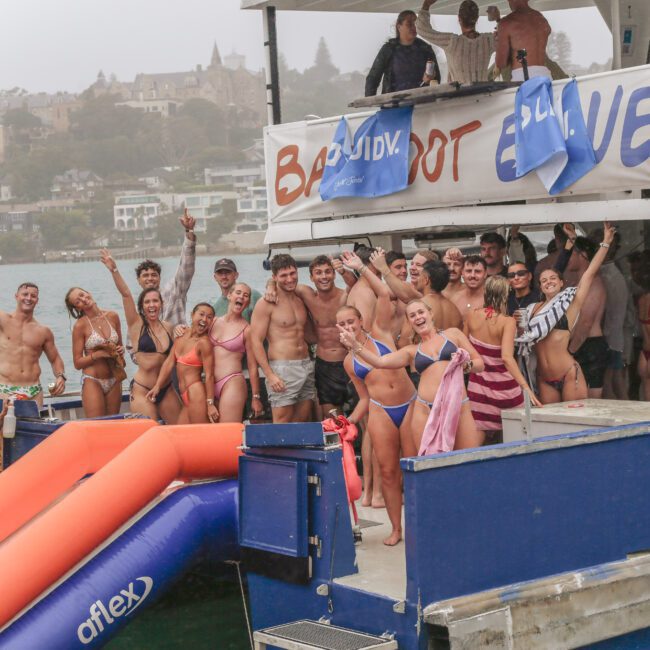 A group of young adults in swimsuits smile and pose on a boat in the water, with banners and an inflatable slide attached. Some people are waving, and houses are visible in the background on a cloudy day.