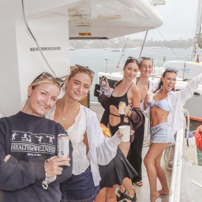 Five young women smiling and holding canned drinks pose together on the deck of a boat. The ocean and another boat are visible in the background on a cloudy day. The scene appears casual and lively.