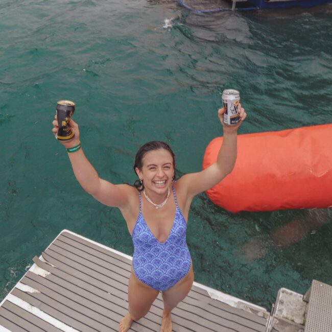 A smiling person in a blue swimsuit stands on a boat ladder, holding up two cans over clear turquoise water, with another swimmer nearby and a large orange float in the background.