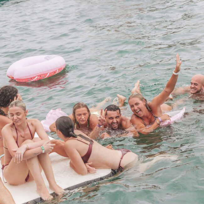 A group of people in swimsuits smile and laugh while relaxing together in the water and on a floating platform, with a pink inflatable ring nearby. The atmosphere is lively and fun.