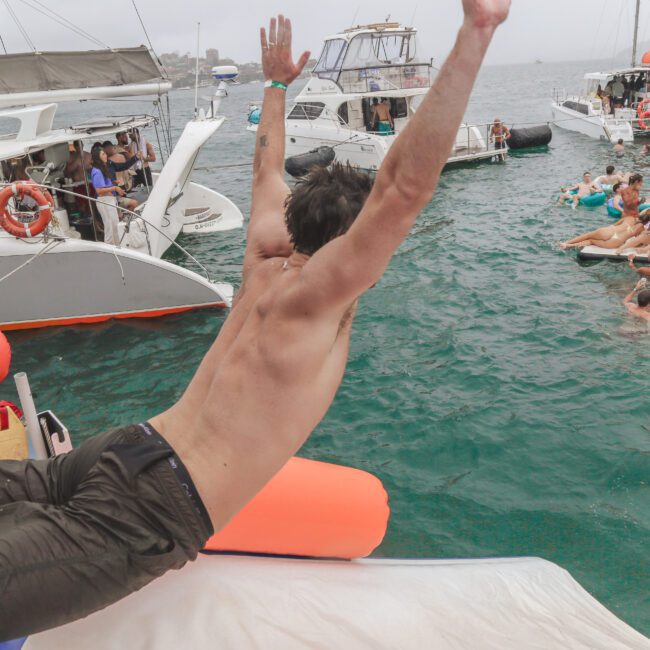 A shirtless man jumps off a yacht into the water, where people are swimming and lounging on other boats nearby during a lively yacht party.
