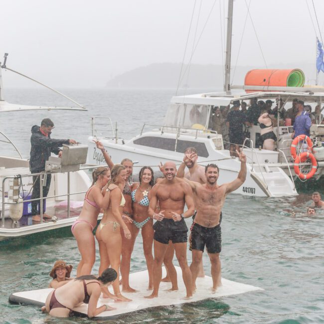 A group of people in swimwear stand and pose on a floating platform in the water, smiling and raising their arms. Other people swim nearby, and two boats with more people are anchored in the background.