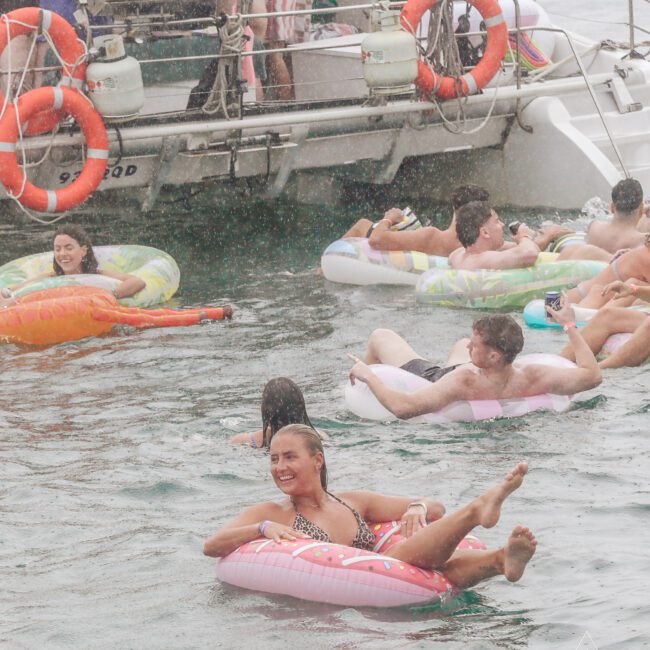 People relax on colorful inflatable tubes in the water near a boat. Some are floating close together, and one woman smiles brightly in the foreground. The scene appears to be a fun social gathering.