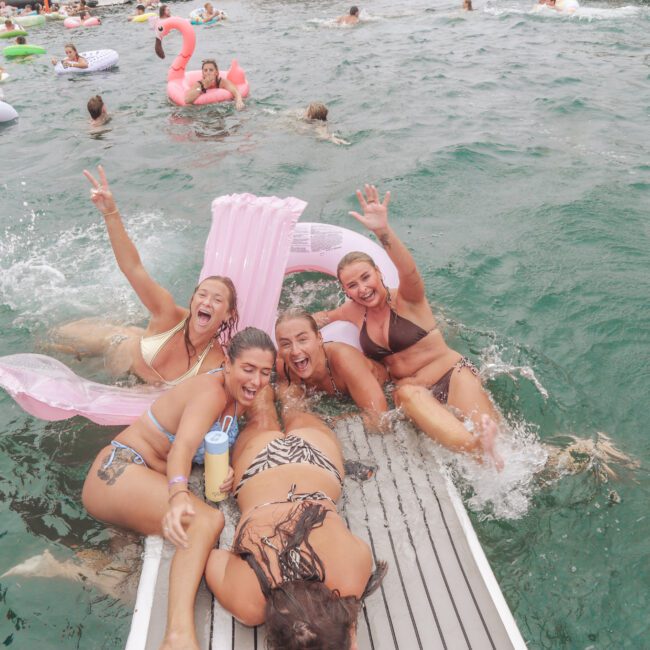 Five women in swimsuits laugh and splash in the water while sitting on a dock, surrounded by people on colorful inflatable floats in a lively, outdoor setting. A pink flamingo float is visible in the background.