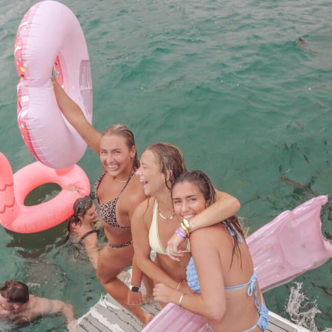 Three young women in swimsuits smile and pose on a dock over green water, holding pool floats. More people are in the water nearby, and there are several inflatable rings and a pink float.