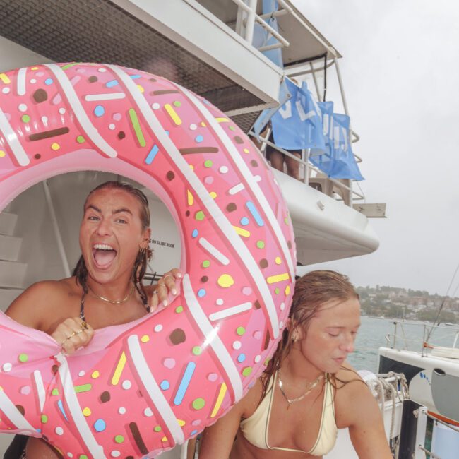 Two women in swimsuits are on a boat. One is smiling and holding a large pink donut-shaped pool float while the other is looking down and adjusting her towel. There are multiple blue chairs and a city shoreline in the background.