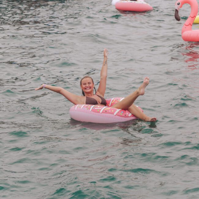 A woman in a pink swimsuit smiles and raises her arms while floating on a pink donut-shaped pool float in the water, surrounded by other colorful inflatables, including a flamingo.