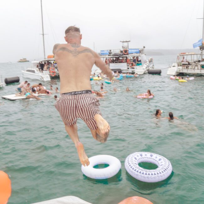 A man in striped swim trunks jumps into the water from a boat, aiming for two floating pool rings. Several people swim and lounge on inflatables nearby, with yachts and boats anchored in the background.