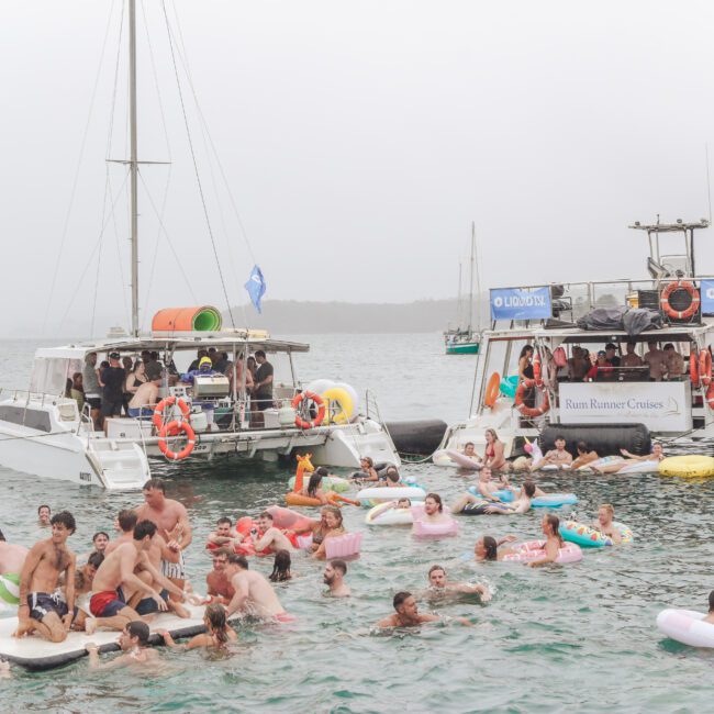 A large group of people swim and relax on colorful floaties near two anchored boats in the ocean, enjoying a lively party atmosphere on a cloudy day.
