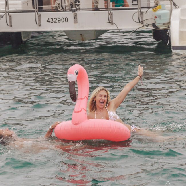 A woman in a white swimsuit floats on a pink flamingo inflatable and smiles, raising one arm, while two people swim nearby in the ocean next to a docked boat.