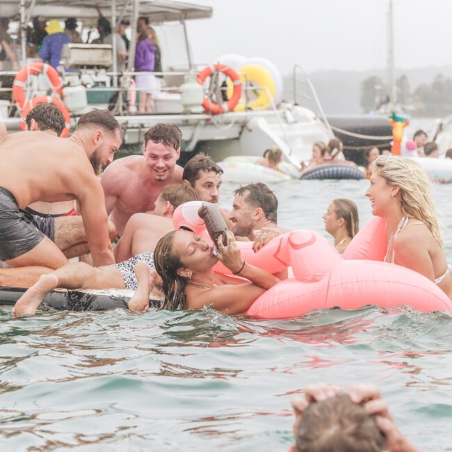 A group of people laugh and play in the water near boats, some climbing onto a floating mat while others float on inflatables, including a large pink flamingo. The scene is lively and joyful.