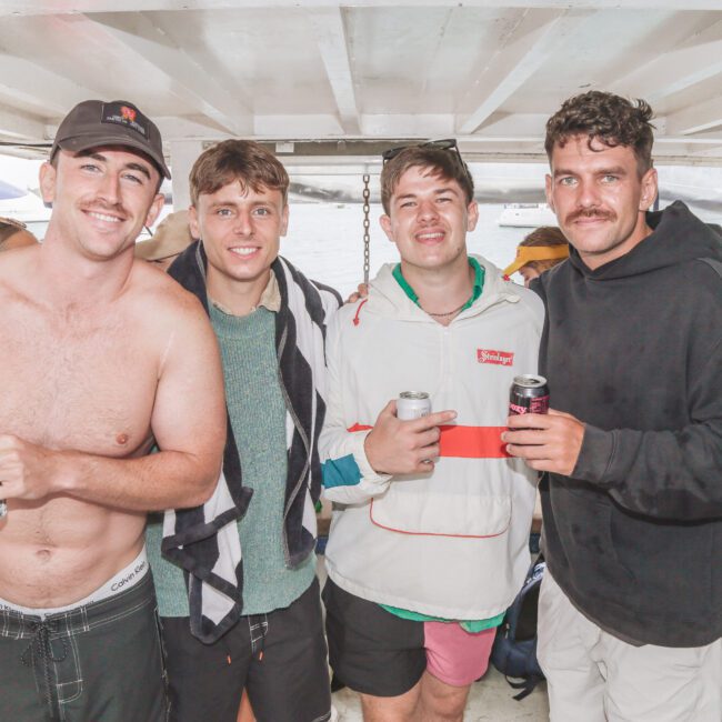 Four young men pose together on a boat, holding drinks and smiling at the camera. The background shows water and parts of other boats. They are dressed casually, some in swimwear and jackets.